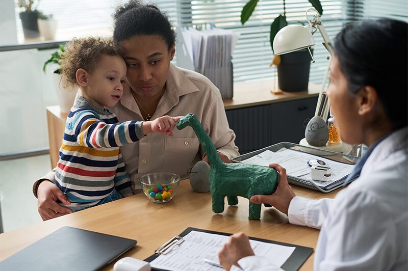 A young child playing with a toy dinosaur while a clinician takes notes nearby.