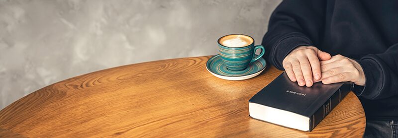 A cup of coffee and a closed book on a wooden table, a hand resting beside them.