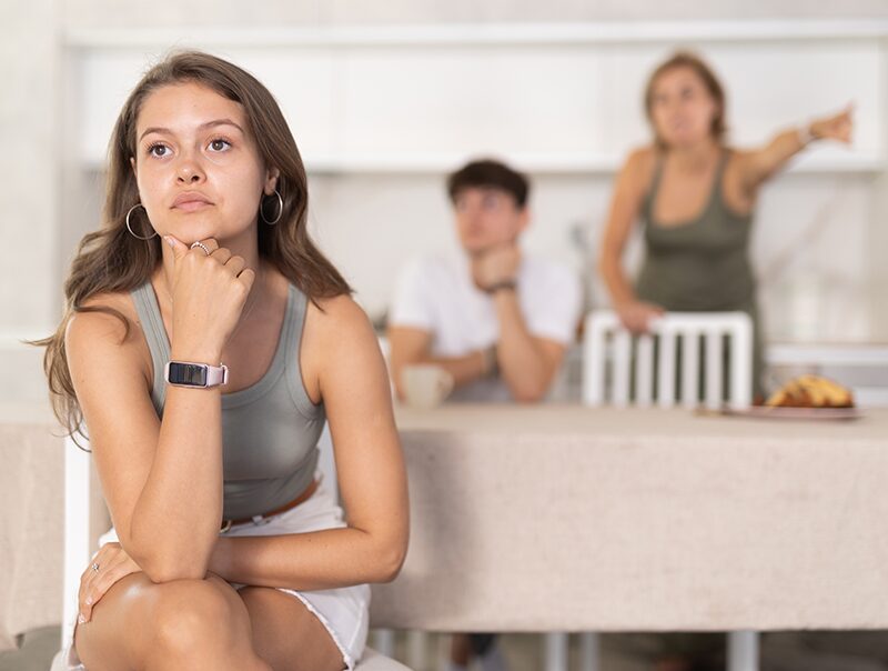 A teenage girl sitting apart at a table while adults argue in the background.